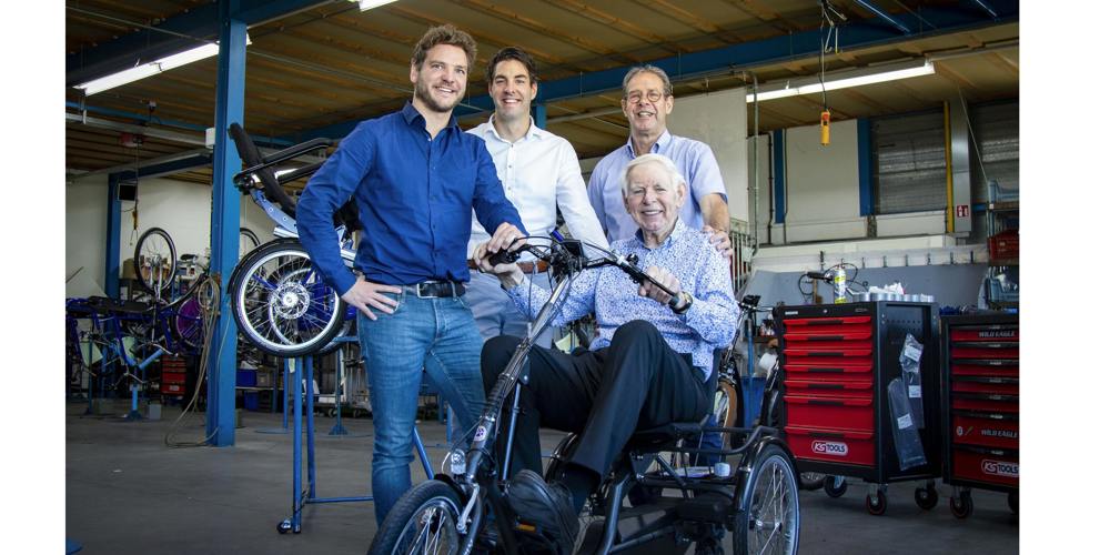 Drie mensen staan om een man die zit op een driewieler in een fietsenmakerswinkel