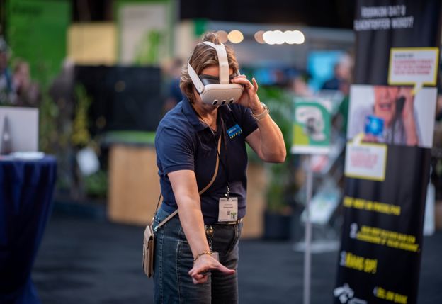Iemand met een VR-bril op haar hoofd bekijkt en beleeft een presentatie tijdens de beurs. Ze probeert iets te pakken dat ze via de VR-bril voor zich ziet.