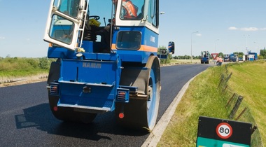 Man in oranje reflecterend hesje in asfaltering auto naast een hectometer paaltje
