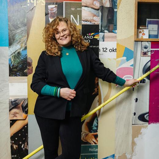 Birte ten Hoopen leunt op gele stang voor kleurrijke muur met posters; ze draagt zwarte blazer en groene trui.