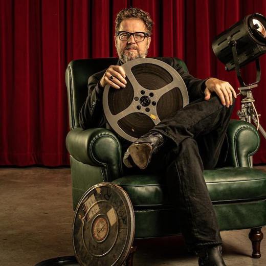 Marin Koolhoven zittend in een groene leren stoel met een filmspoel in zijn handen, voor een rood gordijn op de achtergrond.