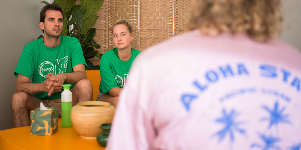 Twee vrijwilligers in een groen shirt van @ease luisteren naar iemand.