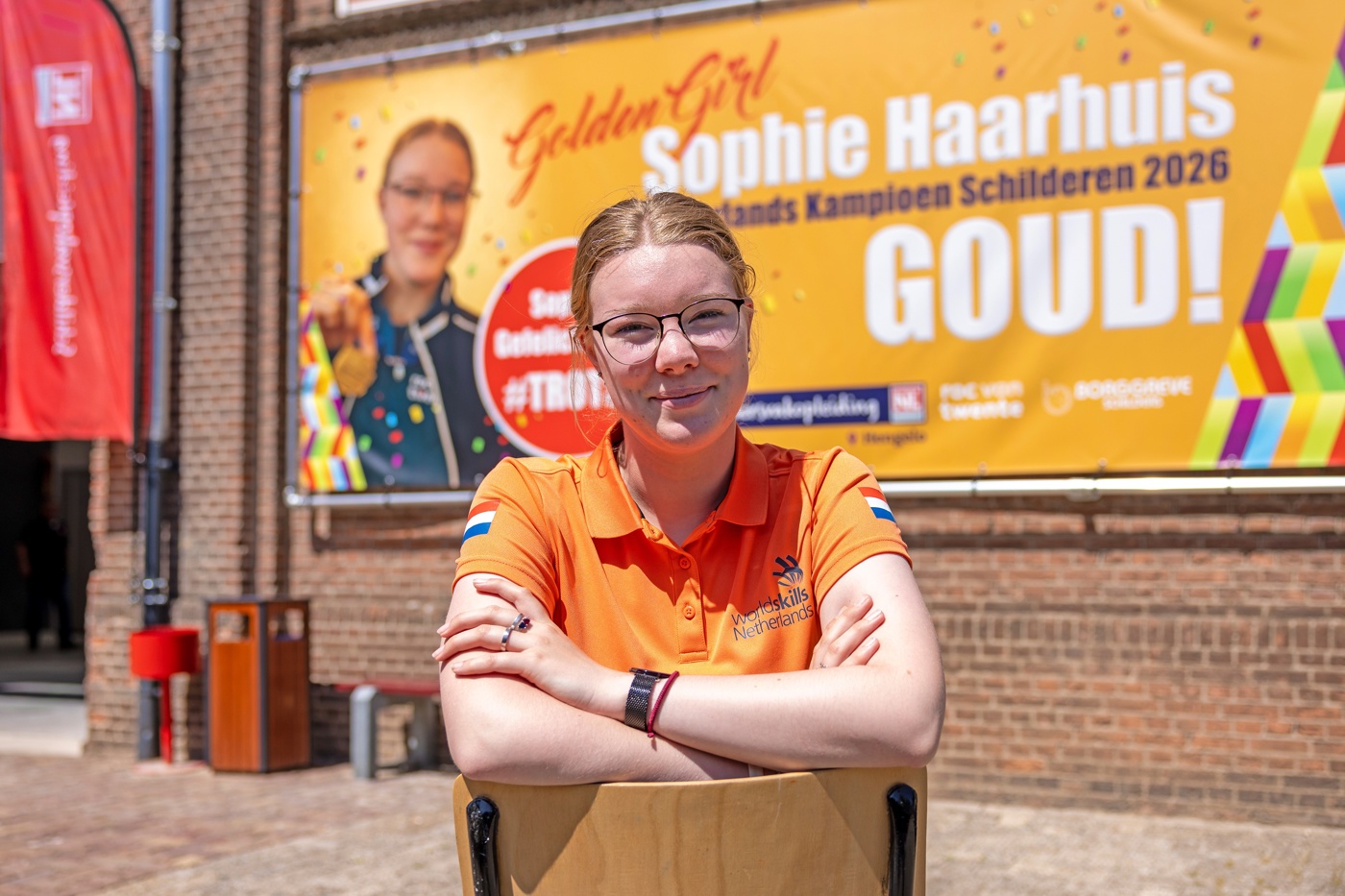 Sophie zit buiten in een oranje shirt op een stoel en kijkt met haar armen over elkaar in de camera. Achter haar hangt een grote oranje banner aan de muur met daarop de tekst: "Golden girl Sophie Haarhuis GOUD!" En de tekst: "Nederlands Kampioen Schilderen 2026". 