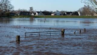 Het beeld toont overstroming in een landelijk gebied, met water dat een hek omringt en huizen in de verte.