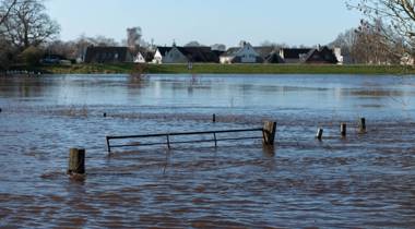 Het beeld toont overstroming in een landelijk gebied, met water dat een hek omringt en huizen in de verte.