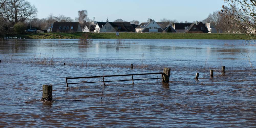 Het beeld toont overstroming in een landelijk gebied, met water dat een hek omringt en huizen in de verte.