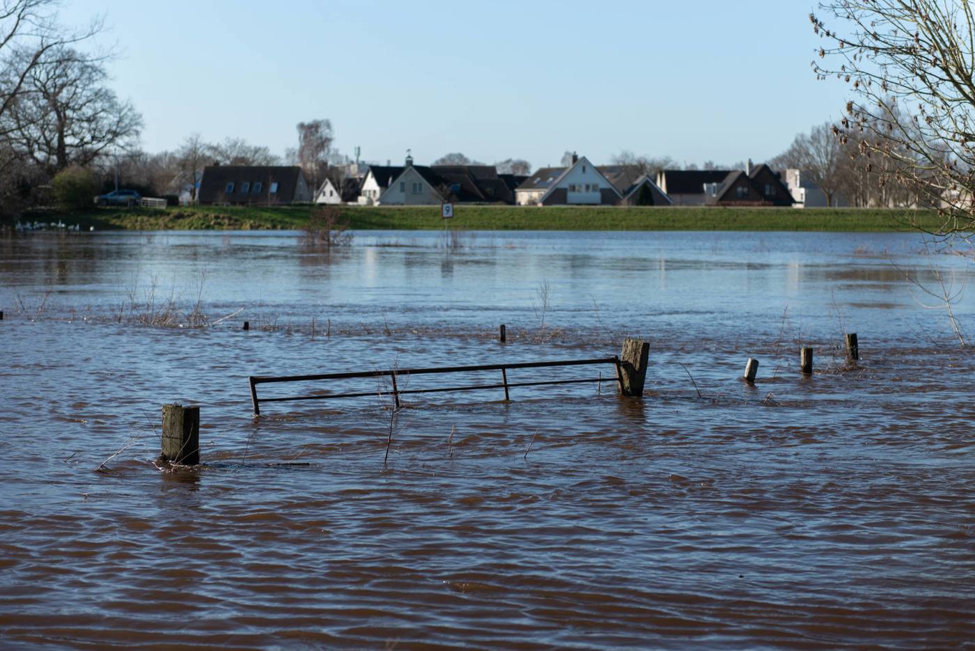 Het beeld toont overstroming in een landelijk gebied, met water dat een hek omringt en huizen in de verte.