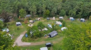 Luchtfoto van een groene camping met caravans, tenten en bomen aan de bosrand.