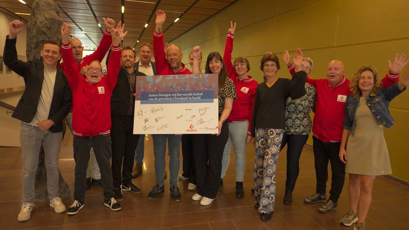 Groep mensen binnen, deels in rode jassen van Special TV, houdt een bord vast met de tekst: "Samen brengen wij het sociale beleid van de provincie Overijssel in beeld", omringd door handtekeningen.