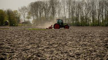 Een tractor ploegt een groot, droog veld met bomen op de achtergrond. (Foto Kenji Elzerman - Unsplash)