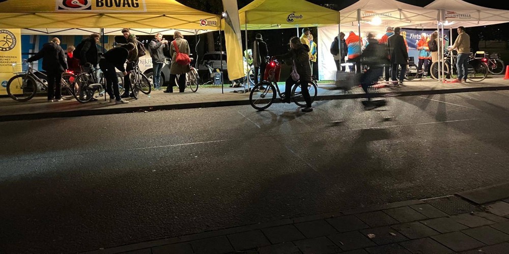 Mensen verzamelen zich bij verlichte tenten van BOVAG en ANWB tijdens een fietsverlichtingsactie in de avond.