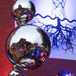 Spiegelende sculptuur met reflectie van festivalpubliek en kunstwerk van stroommast met wortels op bakstenen muur