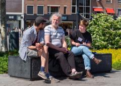 Drie mensen zitten op een bankje in de zon en voeren een gesprek; één persoon maakt aantekeningen.