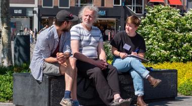 Drie mensen zitten op een bankje in de zon en voeren een gesprek; één persoon maakt aantekeningen.