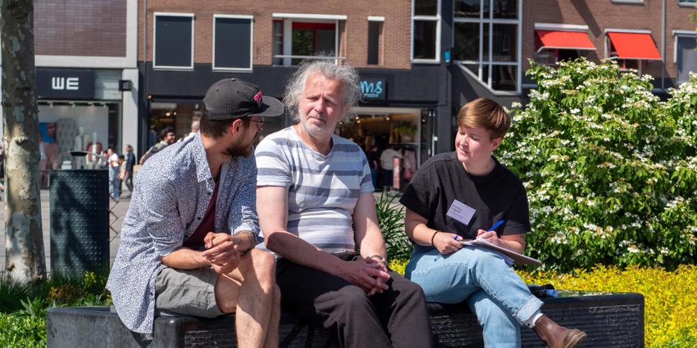 Drie mensen zitten op een bankje in de zon en voeren een gesprek; één persoon maakt aantekeningen.