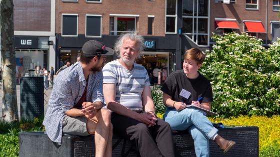 Drie mensen zitten op een bankje in de zon en voeren een gesprek; één persoon maakt aantekeningen.