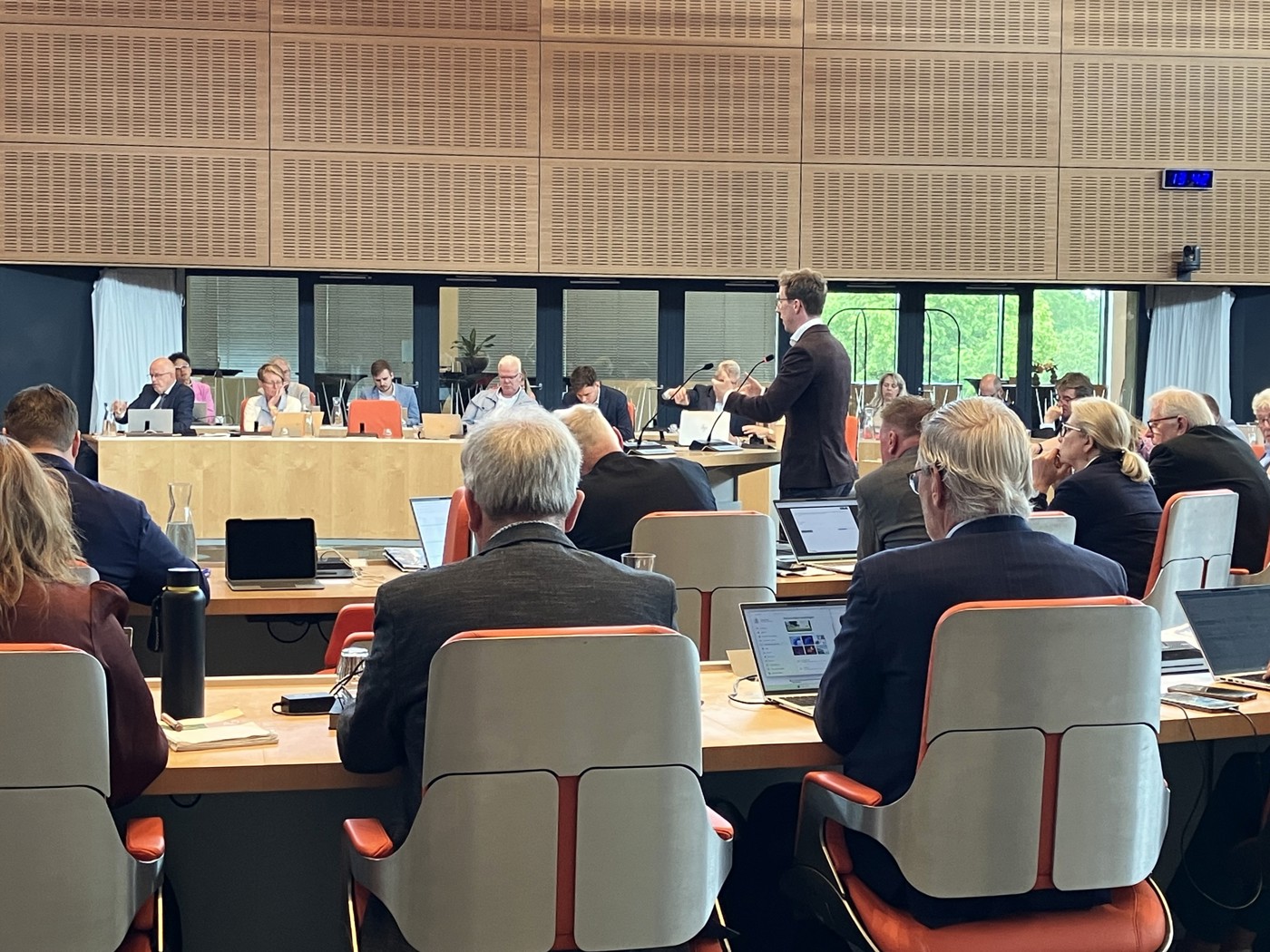 Statenleden aan het werk in de volle Statenzaal. Eén persoon staat bij de interuptiemicrofoon. Op de voorgrond zien we een aantal statenleden op de rug.