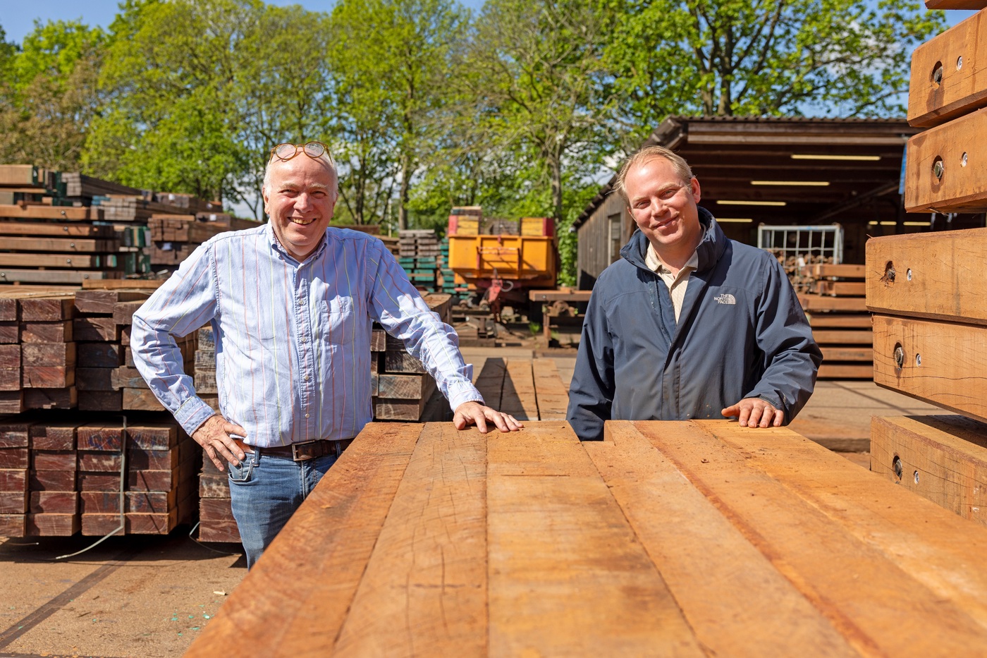 Henk en Jonathan Wesselink op het terrein van het bedrijf achter een grote stapel houten balken en omgeven door nog meer hout.