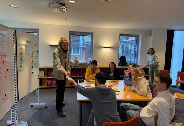 Groep mensen rond een tafel tijdens een workshop, met een staand persoon bij een whiteboard.