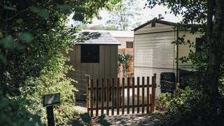 Houten tuinhekje tussen bomen met een schuurtje en stacaravan op de achtergrond.