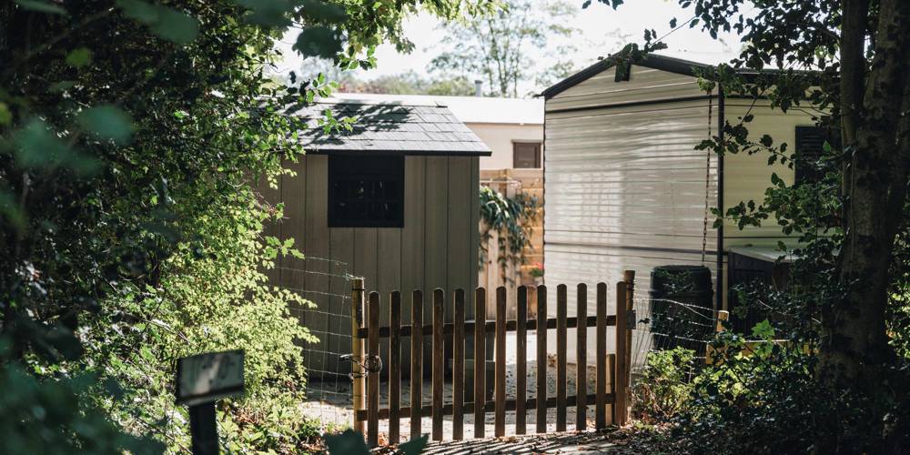 Houten tuinhekje tussen bomen met een schuurtje en stacaravan op de achtergrond.