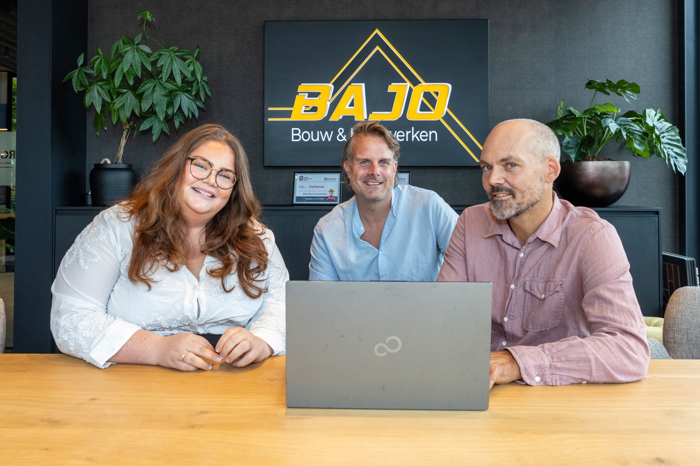 De drie personen op en rij met voor hen een opengeslagen laptop. Achter hen op de muur staat de tekst Bajo Bouw & Bouwwerken.