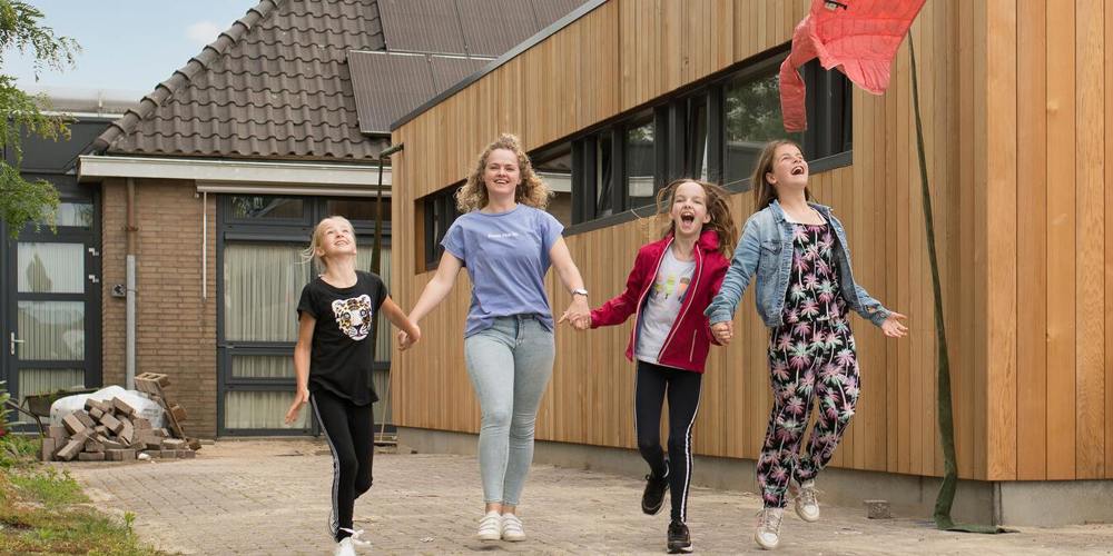 Een jongvolwassene loopt met drie kinderen blij een pad af. Ze houden elkaars hand vast.