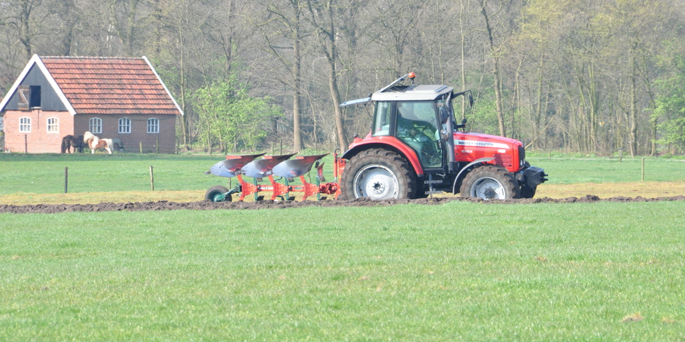 Iemand op de tractor land aan het inzaaien, op de achtergrond een stal en 2 paarden