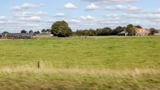 Twee boerderijen gefotografeerd vanuit een rijdend voertuig. Voor de boerderijen liggen grote grasweiden.