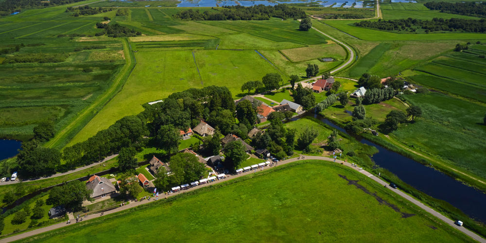 vogel perspectief foto van landschap met bomen en op voorgrond witte kraampjes met mensen op een lange strook weg