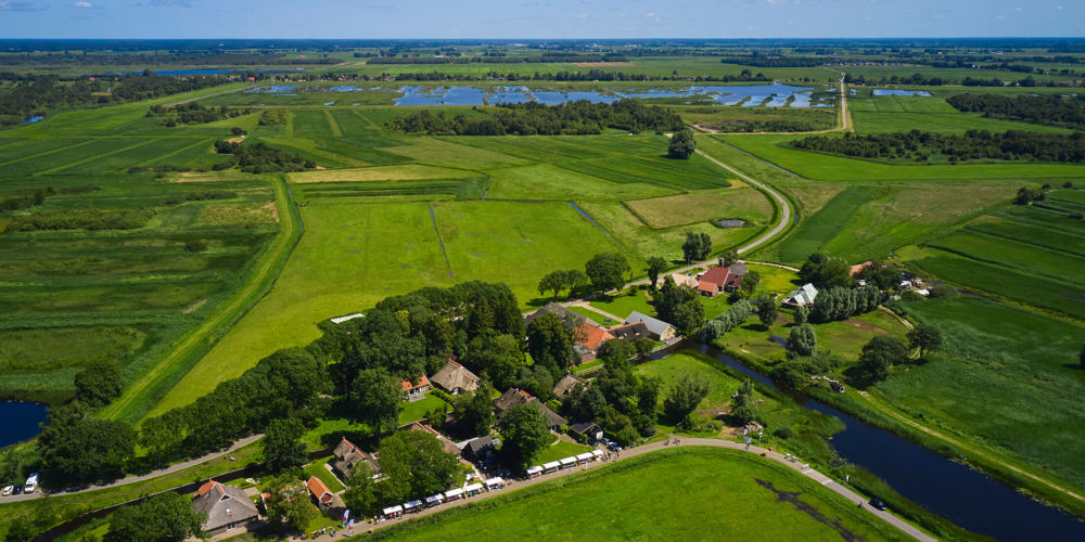 vogel perspectief foto van landschap met bomen en op voorgrond witte kraampjes met mensen op een lange strook weg