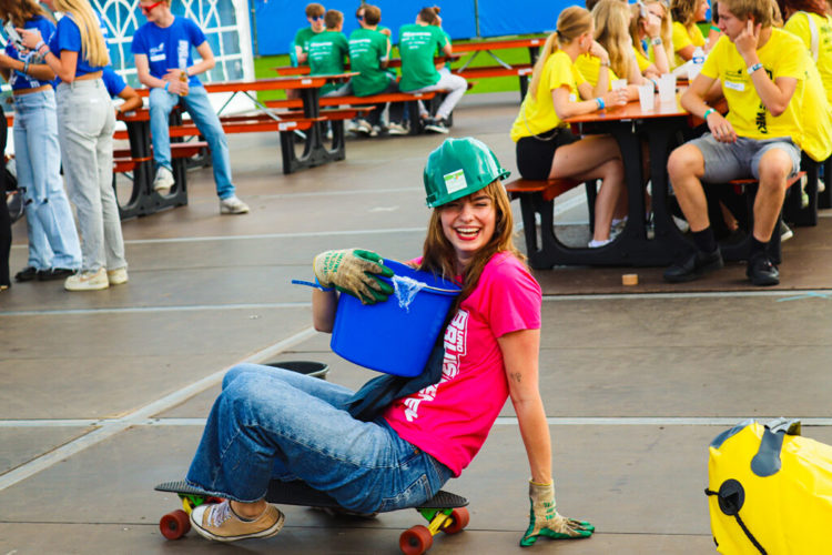Studente zit op een skateboard. Ze houdt een blauwe emmer vast en heeft een groene helm op.