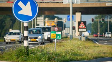 Drukke weg met verkeersborden, waaronder een blauw bord met witte pijl naar links, snelheidslimiet van 80 km/u, en een viaduct.