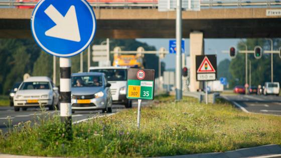 Drukke weg met verkeersborden, waaronder een blauw bord met witte pijl naar links, snelheidslimiet van 80 km/u, en een viaduct.