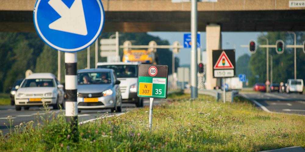 Drukke weg met verkeersborden, waaronder een blauw bord met witte pijl naar links, snelheidslimiet van 80 km/u, en een viaduct.