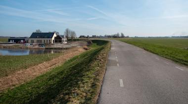 Een geasfalteerde weg loopt langs een dijk, een nieuwbouwhuis en een vijver in een landelijk landschap onder een heldere, blauwe hemel.