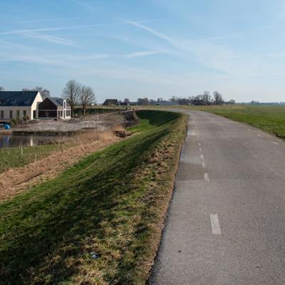 Een geasfalteerde weg loopt langs een dijk, een nieuwbouwhuis en een vijver in een landelijk landschap onder een heldere, blauwe hemel.