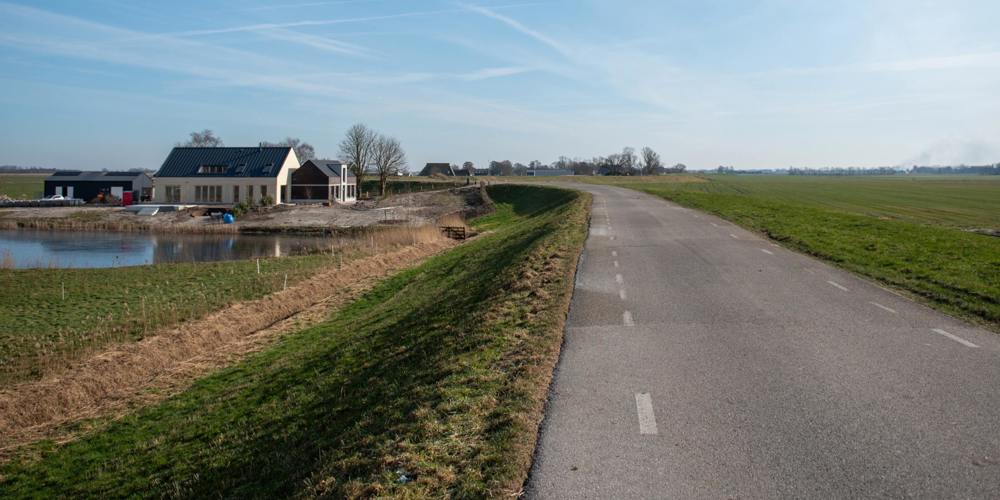 Een geasfalteerde weg loopt langs een dijk, een nieuwbouwhuis en een vijver in een landelijk landschap onder een heldere, blauwe hemel.