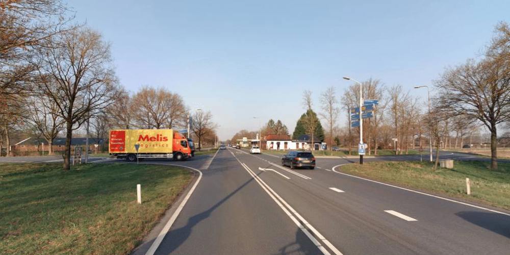 Weg met vrachtwagen en rijdende auto's, omgeven door bomen onder een blauwe lucht.