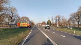 Weg met vrachtwagen en rijdende auto's, omgeven door bomen onder een blauwe lucht.