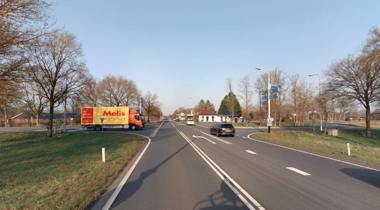 Weg met vrachtwagen en rijdende auto's, omgeven door bomen onder een blauwe lucht.