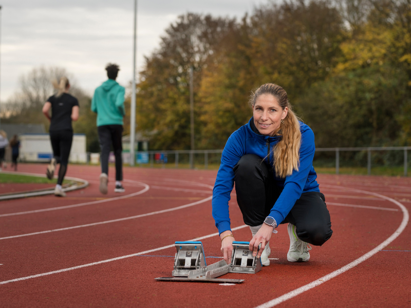 De geïnterviewde in blauwe sportkleding gehurkt bij een startblok op een atletiekbaan. Op de achtergrond zijn hardlopers te zien op de binnenbaan.