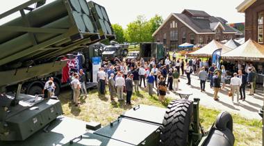 Een buitenevenement met een menigte mensen en militaire voertuigen op een grasveld.