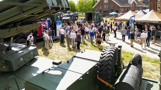 Een buitenevenement met een menigte mensen en militaire voertuigen op een grasveld.