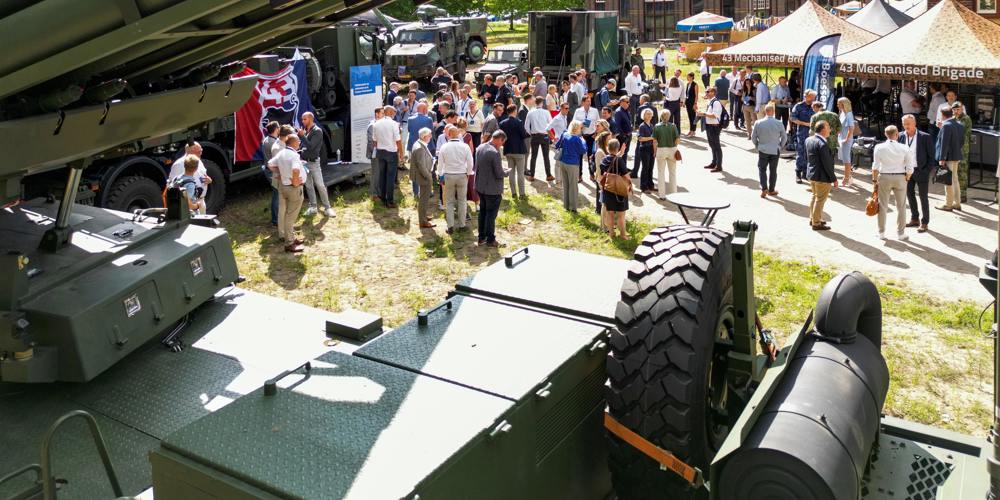 Een buitenevenement met een menigte mensen en militaire voertuigen op een grasveld.