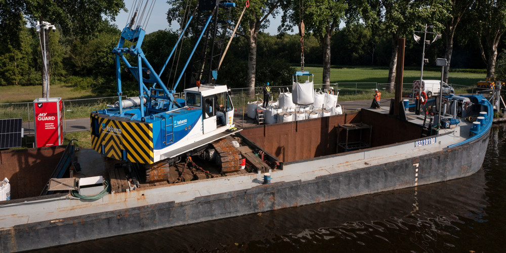 zicht op een boot met kraan die voor de damwand belastingsproef een tweede laag big bag zandzakken plaatst direct langs kanaal Almelo- De Haandrik