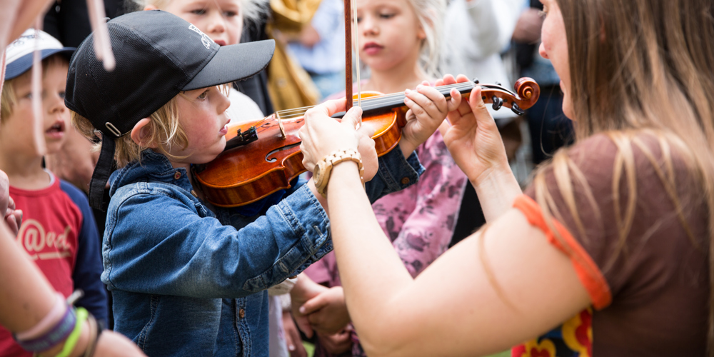 Jongen houd viool vast met behulp van vrouw en een groepje kinderen staat eromheen
