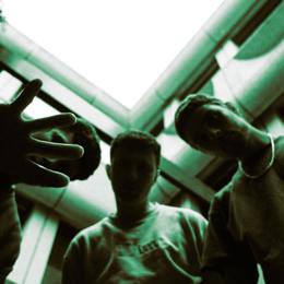 Drie mensen kijken omlaag naar de camera, één met een uitgestrekte hand; gebouw en heldere lucht op de achtergrond, met groene tint