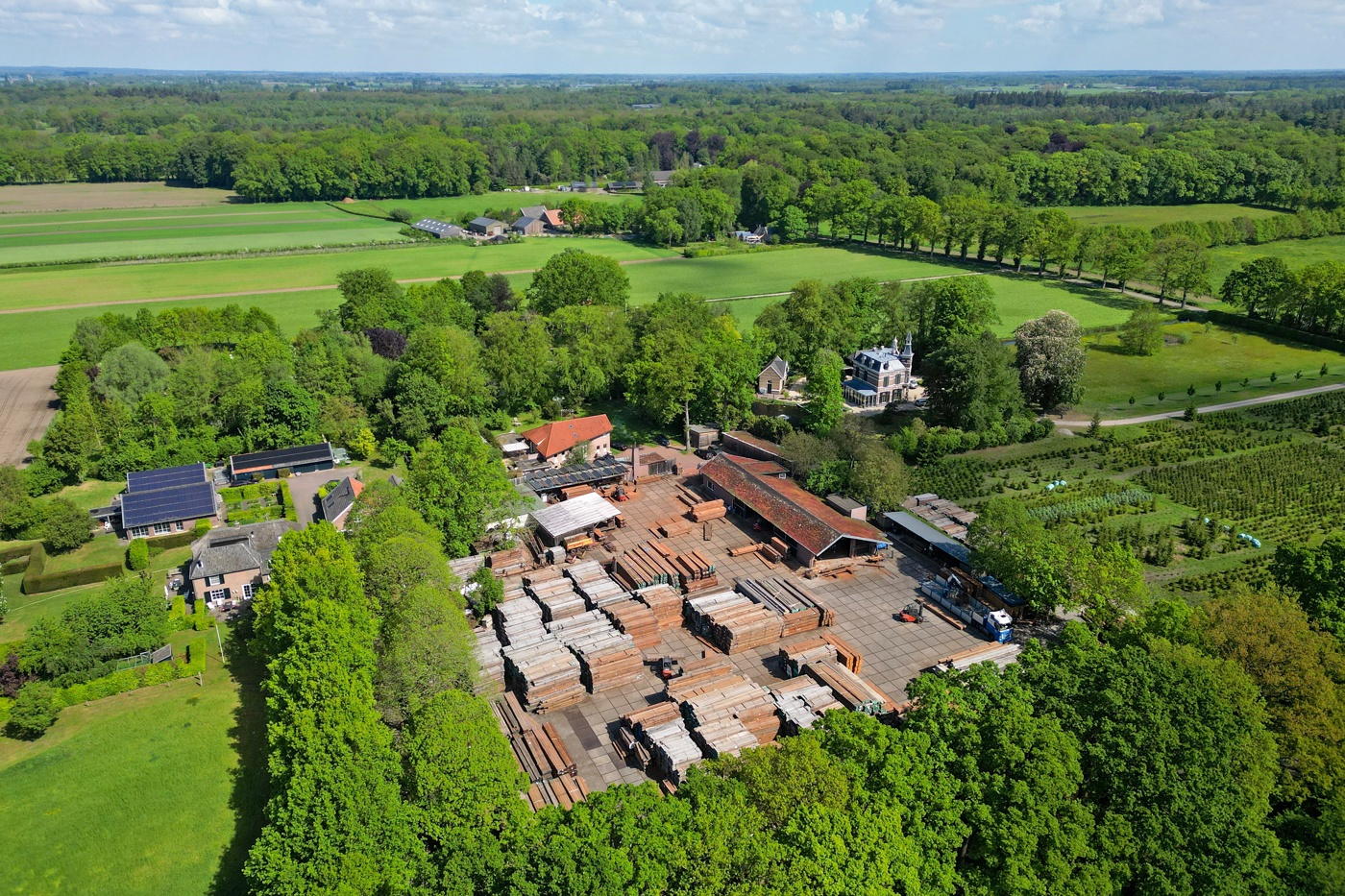 Luchtfoto van het bedrijf waar veel hout ligt opgeslagen, omringd door bomen, weide en bos.