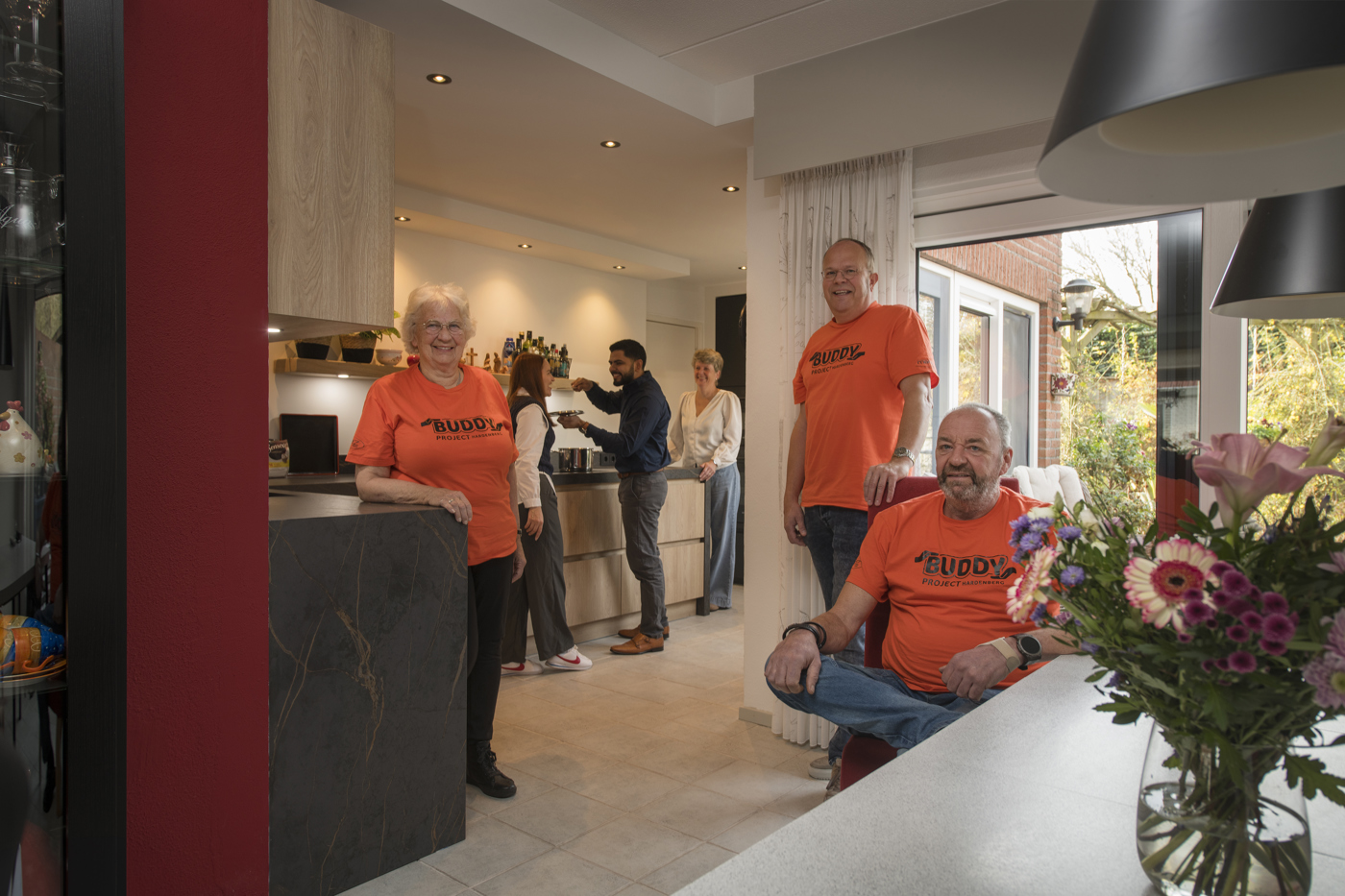 De drie vrijwilligers met een herkenbaar oranje T-shirt met de tekst "Buddy" erop, in een open keuken. Op de achtergrond zijn nog 3 mensen aan het koken en in gesprek in de keuken.